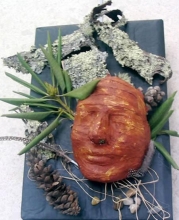 mask surrounded by bark,  pine cones, rocks, feathers, and foliage